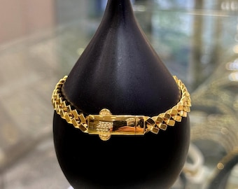 May include: A gold-colored bracelet with a geometric chain design is displayed on a black stand. The bracelet features a rectangular clasp and a series of interconnected, square-shaped links. The jewelry is presented against a blurred background.