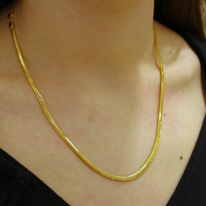 May include: A gold chain necklace with a snake chain design. The chain is thin and delicate, and it is worn by a person with a black shirt.