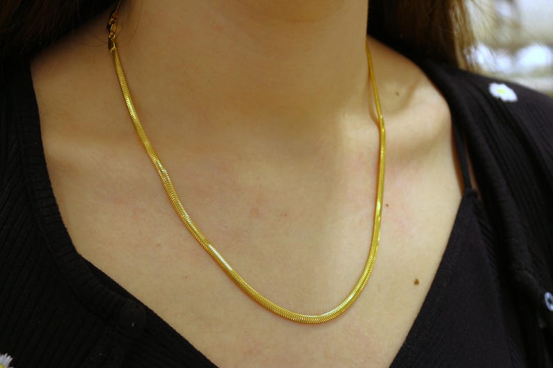 May include: A gold chain necklace with a snake chain design. The chain is thin and delicate, and it is worn by a person with a black shirt.