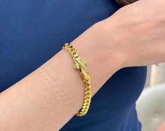 May include: A gold-colored curb chain bracelet with a decorative clasp. The bracelet is worn on a person's wrist, and the links are closely connected. The clasp has an ornate design.