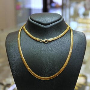 May include: A gold snake chain necklace on a black display stand. The chain is made of small, interlocking links.