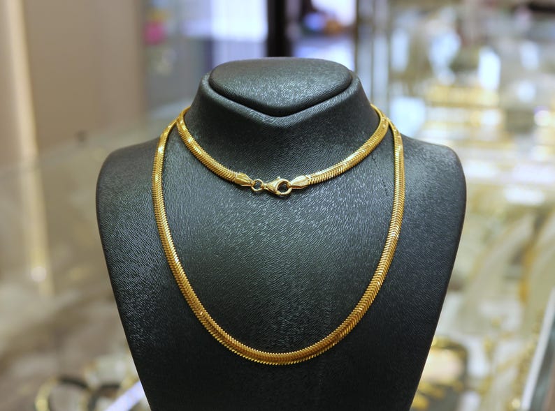 May include: A gold snake chain necklace on a black display stand. The chain is made of small, interlocking links.