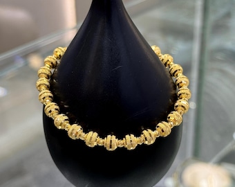 Può includere: Un braccialetto dorato con perline con dettagli strutturati. Il braccialetto è esposto su un supporto nero, mettendo in mostra le sue perline rotonde e il suo design intricato. Il gioiello è di un colore oro brillante.