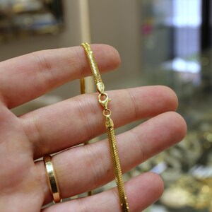 May include: A gold chain necklace with a snake chain design. The chain is held in a hand, and the clasp is visible.