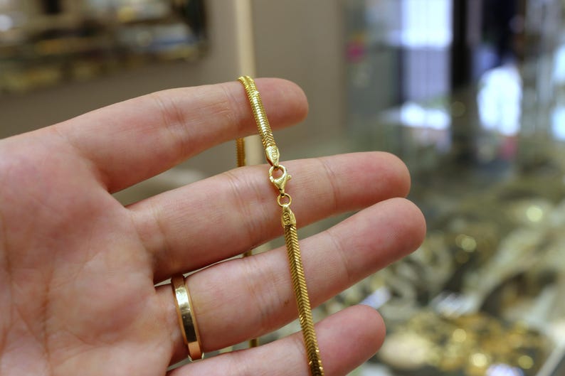 May include: A gold chain necklace with a snake chain design. The chain is held in a hand, and the clasp is visible.