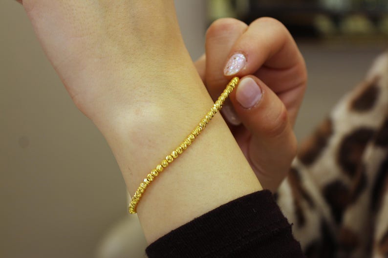 Puede incluir: Un brazalete de oro con un dise&ntilde;o de cuentas. El brazalete est&aacute; en la mu&ntilde;eca de una persona.