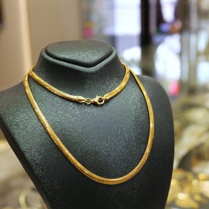 May include: A gold chain necklace on a black display stand. The chain is a snake chain style.