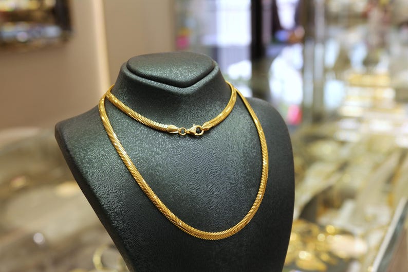 May include: A gold chain necklace on a black display stand. The chain is a snake chain style.