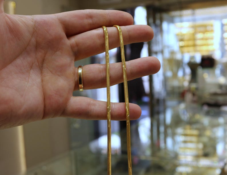 May include: A gold chain necklace with a snake chain design. The chain is held in a hand.