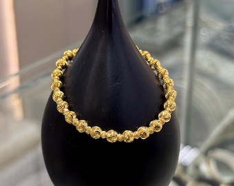 Può includere: Un braccialetto dorato con un design a perline. Il braccialetto è esposto su un supporto nero. Le perline hanno un motivo strutturato e dettagliato. Il gioiello è probabilmente in oro.
