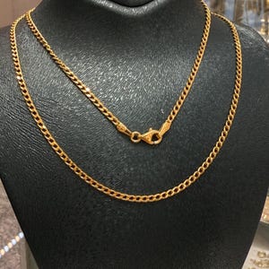 May include: Two gold-colored chain necklaces displayed on a black surface. The necklaces have a curb chain design, with a clasp visible on the upper necklace. The chains are likely made of gold or a gold-colored metal.