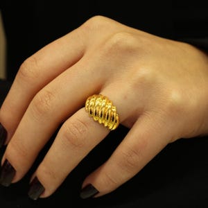 May include: A gold-colored ring with a textured, ridged design. The ring is worn on a finger, showcasing its intricate details and bright, reflective surface. The jewelry piece is a statement accessory.