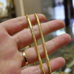 May include: Two gold chains, one slightly thicker than the other, are held in a hand. The chains are both snake chains, with a smooth, shiny finish.