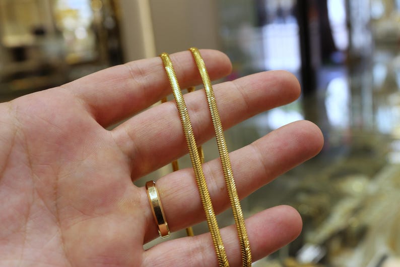 May include: Two gold chains, one slightly thicker than the other, are held in a hand. The chains are both snake chains, with a smooth, shiny finish.
