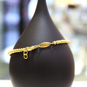 Puede incluir: Un brazalete de cuentas doradas con una peque&ntilde;a etiqueta dorada que dice "Melissa Joy". El brazalete se muestra en un soporte negro.