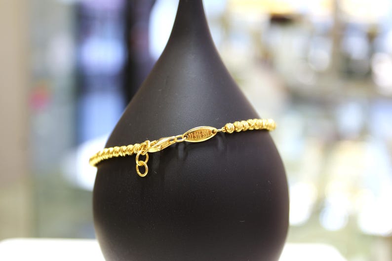 Puede incluir: Un brazalete de cuentas doradas con una peque&ntilde;a etiqueta dorada que dice "Melissa Joy". El brazalete se muestra en un soporte negro.