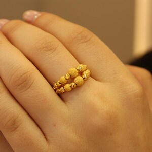 May include: A gold ring with a textured design featuring multiple small round beads. The ring is worn on the middle finger of a hand.
