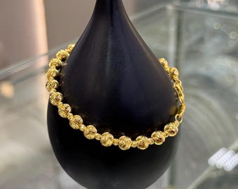 Può includere: Un braccialetto d'oro con una serie di perline sferiche strutturate. Il braccialetto è esposto su un supporto nero a forma di goccia. La chiusura è visibile e il braccialetto sembra essere fatto di un metallo giallo lucido.