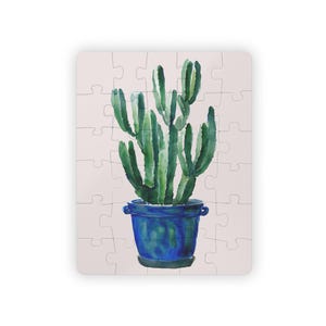 May include: A watercolor puzzle featuring a green cactus in a blue pot. The cactus has multiple upright stems. The pot is a deep blue with a textured appearance. The puzzle pieces are a light beige color.
