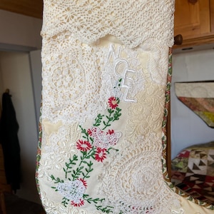 May include: Cream-colored Christmas stocking with intricate lace and embroidered floral details. The stocking features the word "NOEL" in white letters and is adorned with red flowers and green leaves. The top is trimmed with a delicate lace.