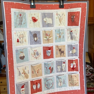 Op de afbeelding: Een gewatteerde kerst adventskalender met 25 genummerde vakken, elk met een unieke illustratie met vakantiethema. De kalender heeft een rode rand met een witte en grijze achterkant en wordt aan een hanger gehangen.
