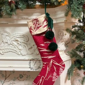 Puede incluir: Calcetín de Navidad rojo y blanco con un diseño de escena de invierno. El calcetín tiene un puño de terciopelo verde y tres pompones verdes.