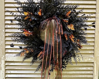 May include: A Halloween wreath with black foliage, orange accents, and a sheer ribbon with orange stripes. A small white ghost decoration hangs above the wreath. The wreath is displayed on a weathered, cream-colored shutter.