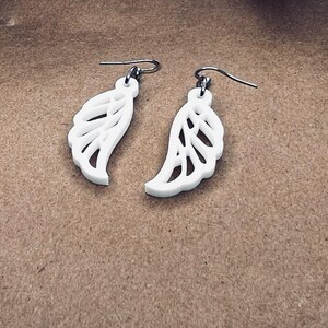 May include: A pair of white dangle earrings with a delicate, intricate wing design. The earrings are made of a lightweight material and have a silver-toned hook.