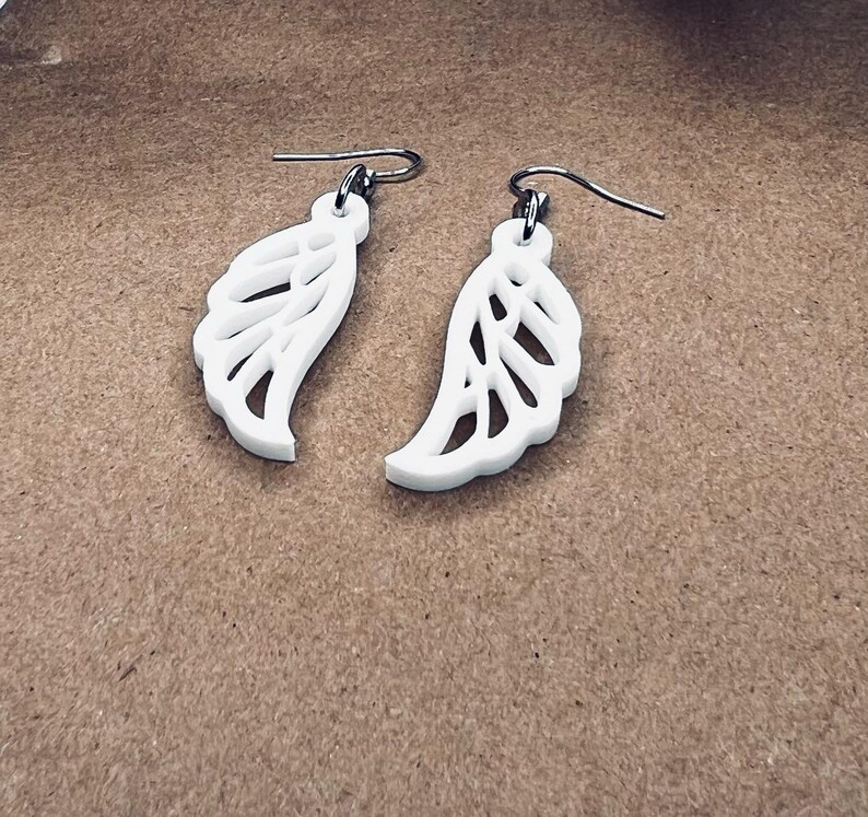 May include: A pair of white dangle earrings with a delicate, intricate wing design. The earrings are made of a lightweight material and have a silver-toned hook.