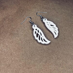 May include: A pair of white dangle earrings with a delicate wing design. The earrings are made of a lightweight material and have a silver hook.