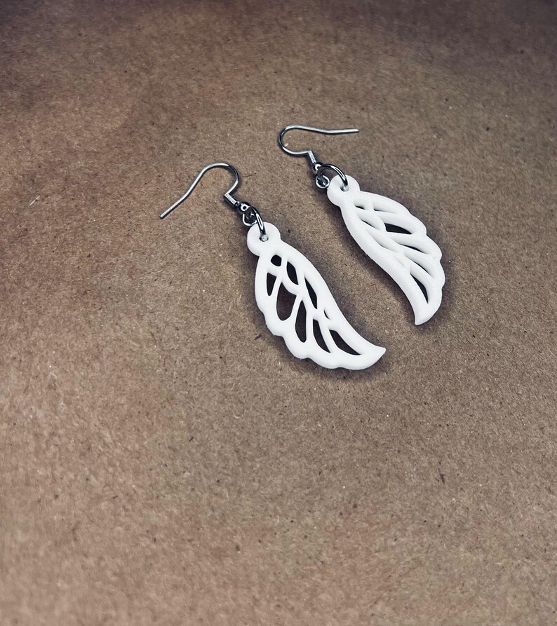 May include: A pair of white dangle earrings with a delicate wing design. The earrings are made of a lightweight material and have a silver hook.