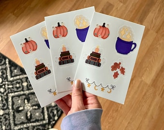 May include: Three white sticker sheets featuring fall-themed illustrations. Each sheet has a pumpkin, a stack of books with a candle, and a blue mug with sparkles. The stickers are held in a person's hand.