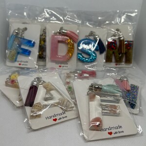May include: A selection of handmade keyrings in clear packaging. The keyrings display letters, seashells, and other decorative elements. Some keyrings have colourful tassels. The cards read "Handmade with love."