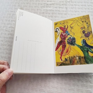 May include: A postcard featuring a colorful painting of a red bull playing a musical instrument with a woman in a white dress and a woman in a green dress. The background is yellow with a blue sky.