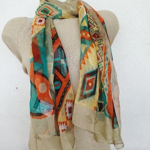 Può includere: Una sciarpa colorata con motivi geometrici in tonalità arancio, blu, verde e giallo. La sciarpa è realizzata in tessuto trasparente e ha un bordo a frange.