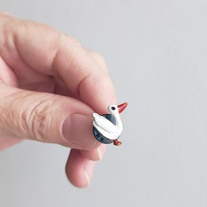 Può includere: Una piccola spilla decorativa raffigurante un cigno bianco con becco e zampe rosse. Il cigno è posato su una base blu scuro con un bordo argentato. La spilla è tenuta in mano.