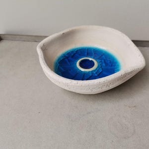 Puede incluir: Un cuenco de cerámica blanco con un diseño de ojo azul. El cuenco tiene un exterior texturizado y un interior liso y brillante con un patrón de mosaico azul y un centro blanco. El cuenco está sobre una superficie gris claro.