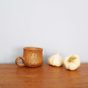 Puede incluir: Una taza de cerámica marrón con textura rugosa se encuentra sobre una superficie de madera junto a dos bulbos de ajo enteros.