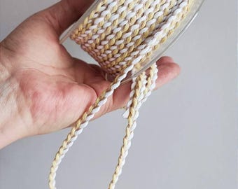 Puede incluir: Un carrete de cordón trenzado blanco y beige. El cordón está trenzado y tiene un brillo sutil.
