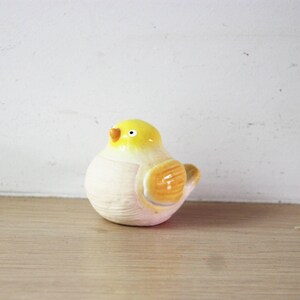May include: A ceramic bird figurine with a yellow head and orange beak and wings. The bird has a white body and is sitting on a wooden surface. The background is a white wall.