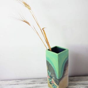 Puede incluir: Un jarrón rectangular alto con un esmalte verde, azul y beige. El jarrón tiene una abertura cuadrada en la parte superior y está decorado con un diseño floral. El jarrón está lleno de tallos de trigo secos.