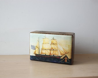 May include: A wooden box with a painted image of a sailboat with a Greek flag on the front. The sailboat has white sails and is sailing on a blue sea. A woman is swimming in the water behind the boat.