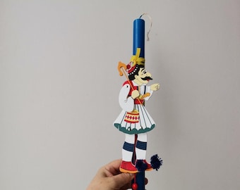 May include: A blue and white wooden Easter candle with a painted figure of a man wearing a traditional Greek costume. The figure is holding a stick and a drum. The candle is decorated with blue and white pom-poms.