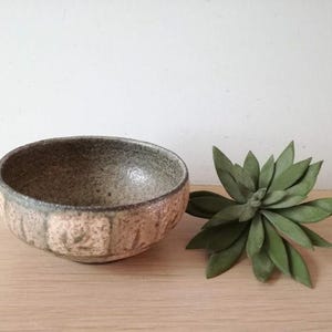 May include: A small, round, ceramic bowl with a light brown and green glaze. The bowl has a textured surface and is slightly wider at the top than the bottom.