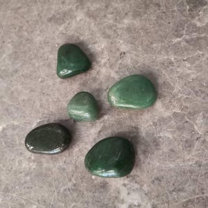 May include: Five smooth, green, polished stones of various sizes and shapes on a gray background.