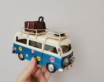 May include: A blue and white toy van with a luggage rack and a brown suitcase on top. The van has pink and yellow flowers painted on the side.