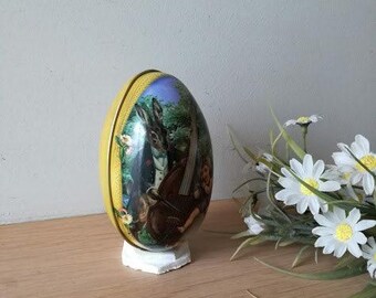 May include: A yellow and blue egg-shaped tin with a picture of a rabbit playing a cello with a girl in a blue dress. The tin is sitting on a wooden surface.
