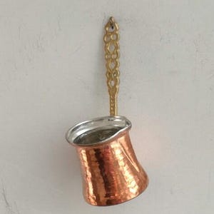 Può includere: Una caffettiera di rame con finitura martellata e manico dorato. La caffettiera è appesa a un gancio su una parete bianca.