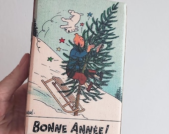 May include: Vintage rectangular box featuring a cartoon illustration of a man on a sled, holding a Christmas tree, with a dog in the air. The text "Bonne Année!" is at the bottom. The box has a light blue background.