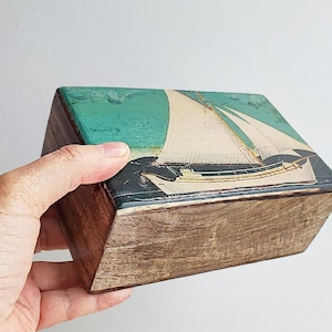 Könnte beinhalten: Eine Holzkiste mit einem Segelbootbild auf dem Deckel. Das Segelboot ist weiß mit grünem und blauem Hintergrund. Die Kiste ist aus dunklem Holz.